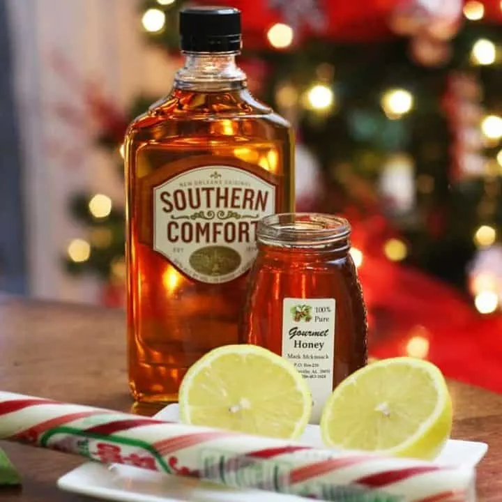 Bottle of Southern Comfort, a jar of honey, a halved lemon, and a peppermint stick.
