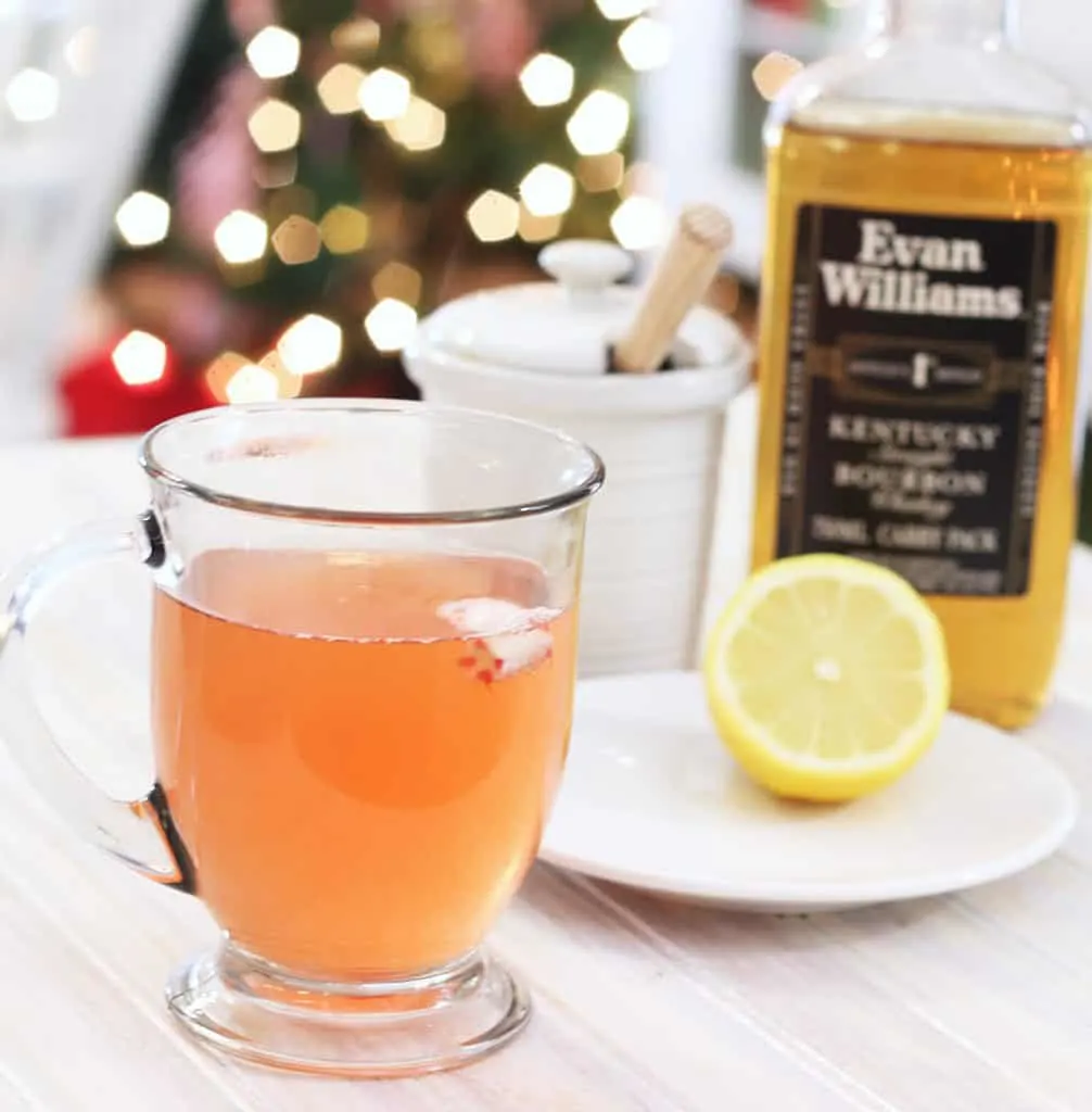 A glass cup with peppermint whiskey honey and lemon on a white tray with a bottle of whiskey and half cut lemon on a plate.