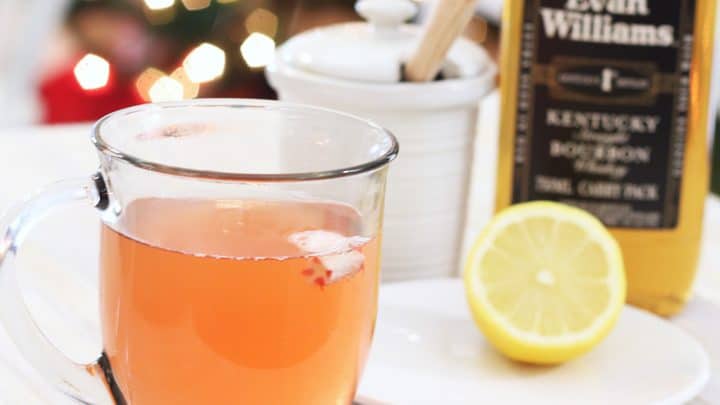 A glass cup with peppermint whiskey honey and lemon on a white tray with a bottle of whiskey and half cut lemon on a plate.
